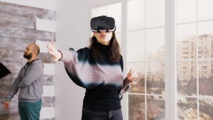 People are making virtual tours of real estate. They are in the empty apartment. A woman is using vr google, while real estate agent is talking with another client in the background
