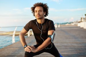 A male afro-american jogger that is stretching legs before running wears a smartphone attached to his arms with a fitness app