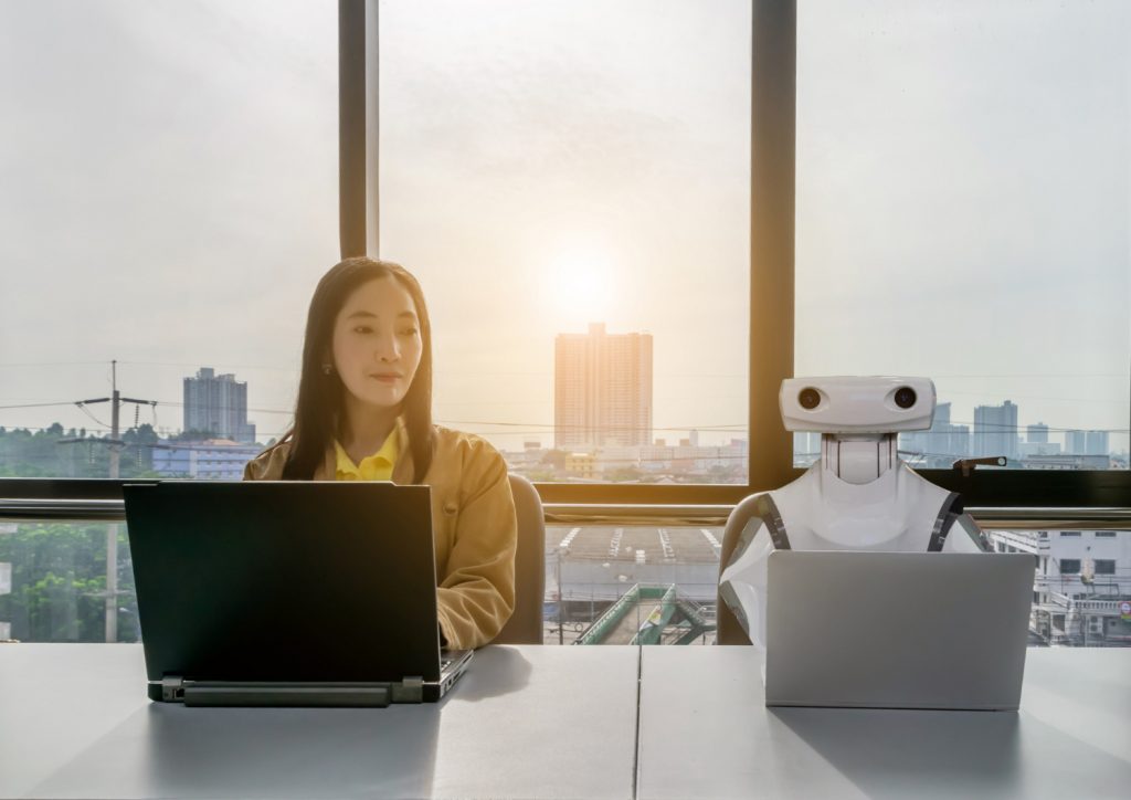 A young Asian women sits in front of computers in the office. A robot is seated next to her to exemplify the use of rpa in real estate. Window view in the background shows skypcrapers.