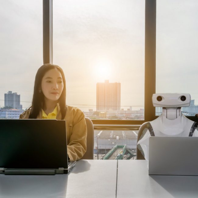 A young Asian women sits in front of computers in the office. A robot is seated next to her to exemplify the use of rpa in real estate. Window view in the background shows skypcrapers.